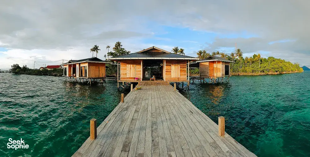 Wooden stilt sea lodges on a tropical island coastline, with a walkway leading out over clear water in Semporna