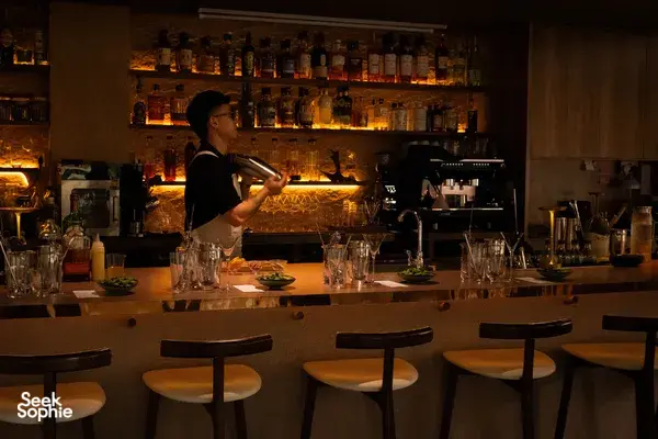 Warm-lit cocktail bar in Singapore with a bartender behind the counter, bottles on shelves, and glasses waiting for drinks