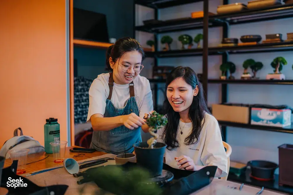 Two participants in aprons working on a tropical bonsai together at a Japanese-style workshop in Singapore, surrounded by tools
