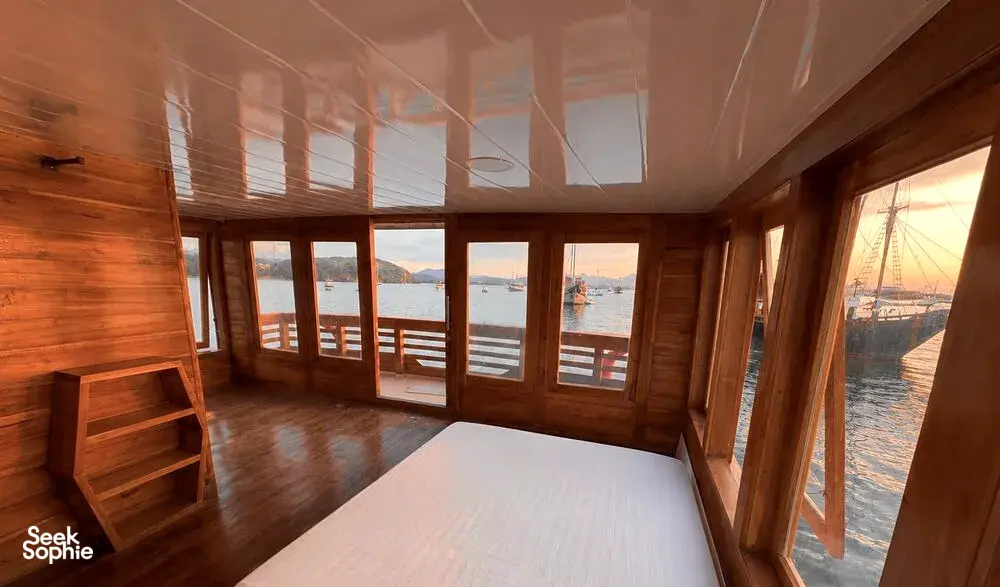 Wooden phinisi lounge with windowed walls and a view over the Komodo waters on a shared 3D2N cruise from Labuan Bajo