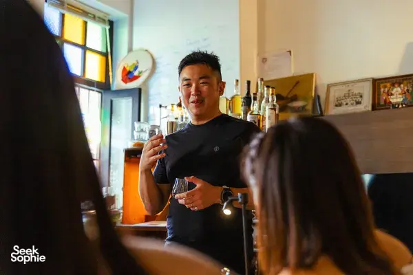 Host explaining flavours during a small-batch whisky tasting in Singapore, reaching up a bottle as guests listen in a home bar