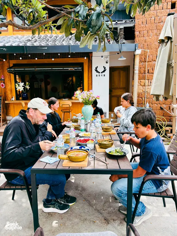 Group enjoying lunch outdoors in Lijiang at a brick courtyard table during a shared Yunnan cooking experience, with steaming bowls