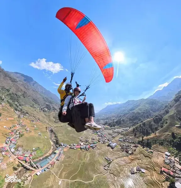 Two paragliders floating over patchwork terraces and a winding river, red-orange canopy catching the wind above the mountains in Sapa