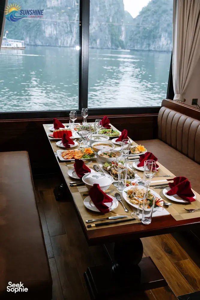 Sunshine Golden Cruises luxe dining table set on a Cat Ba 2D1N overnight cruise, with bay and limestone cliffs beyond the window