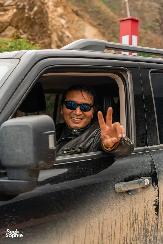 Tibetan guide smiling and making a peace sign from a jeep window on the road during the 6-day Shangri-La Loop