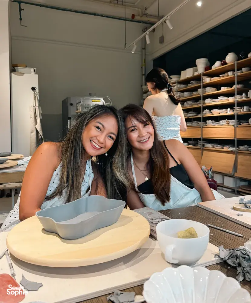 Two friends smiling at a Singapore pottery studio table with European-style ceramic plates and bowls drying behind them