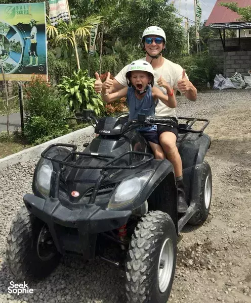 Rider smiling and giving thumbs-up on a black ATV quad bike in Bali, with a child seated in front