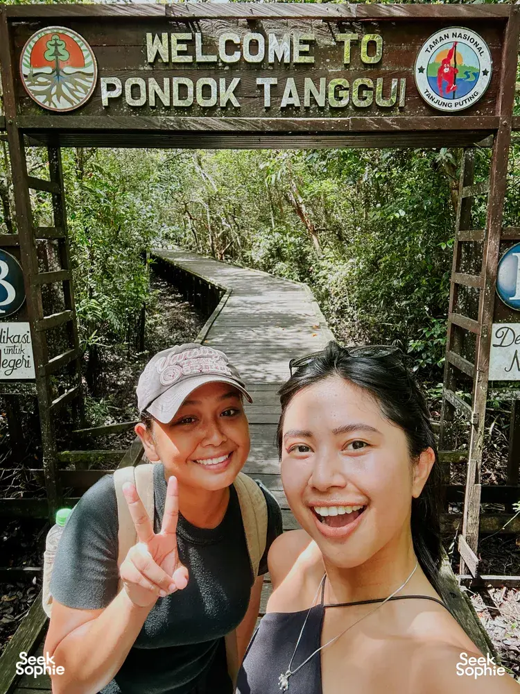 Smiling travellers posing with peace signs by a welcome gate sign for Tanjung Puting National Park, on the boardwalk