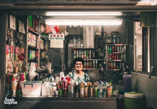 Chef in a Singapore hawker stall serving from behind a counter lined with jars and tins, with a “self-service” sign