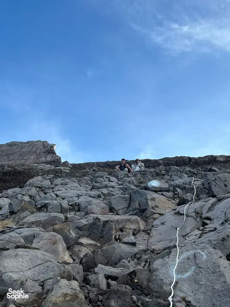 Hikers crouch on Mount Agung’s rocky summit ridge under a bright blue dawn sky during a private sunrise trek with local guides