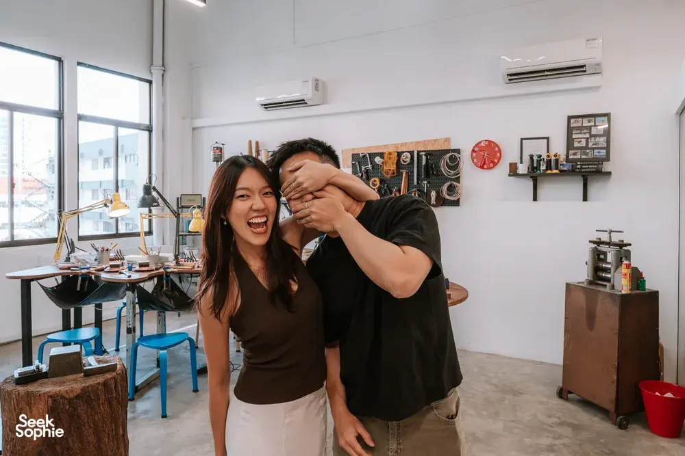Laughing couple posing in a bright silversmithing studio with workbenches and tools behind them at Fat Anvil Studios, Singapore