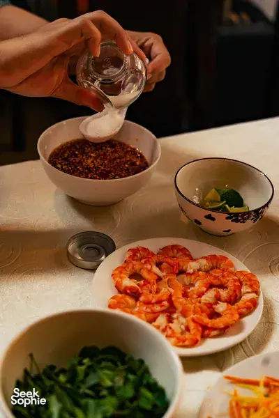 会安 Jolie 烹饪课：双手将香料酱汁倒入碗中，旁边摆着装盘好的虾和新鲜香草，呈现越南家常料理