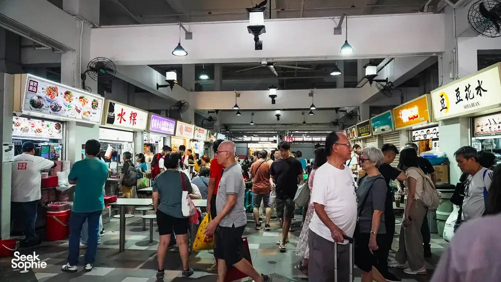 People queueing beneath hawker-centre signs in Singapore on a host-led tour sharing hidden hawker stories with a Nat Geo researcher chef