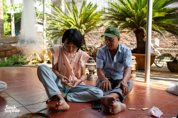 Participants sit and knot pieces of fishing net with an older guide in a shaded courtyard before a Hoi An river journey
