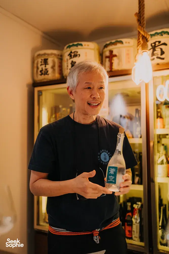 SakeMama smiling and holding a bottle for rare sake tasting in a tucked-away Singapore shop with other brews — Singapore hidden gems