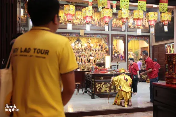 Hey Lad Tour guide in a yellow shirt stands near an ornate altar display with offerings beside shelves of statuettes in Singapore