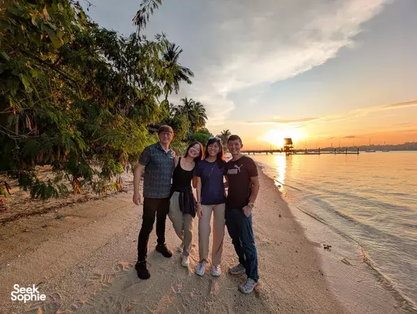 Friends posing on a sandy beach at sunset with calm water and low boats in the distance near Singapore
