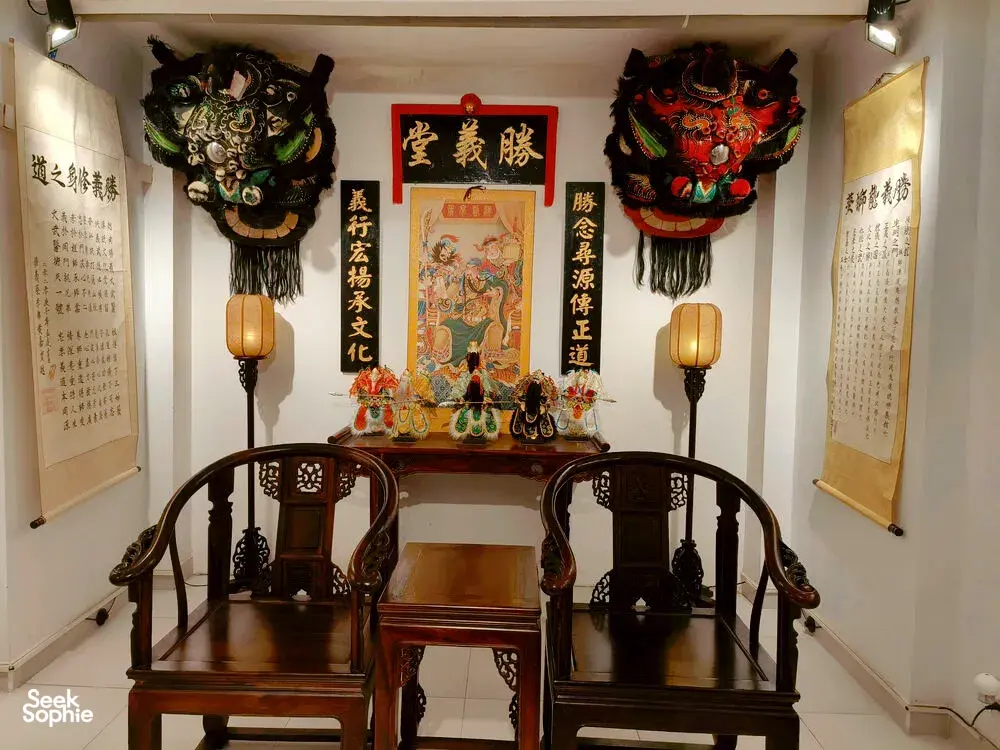 Temple room with two ornate lion dance masks above lanterns, Chinese posters on the walls, and a ceremonial prayer table in Ho Chi Minh City