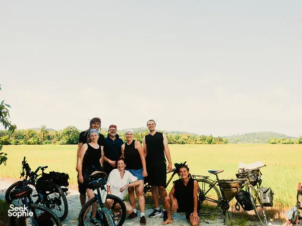 Bicycle Day Langkawi group photo in a sunlit field on Langkawi, posing with bicycles and camping gear by the roadside