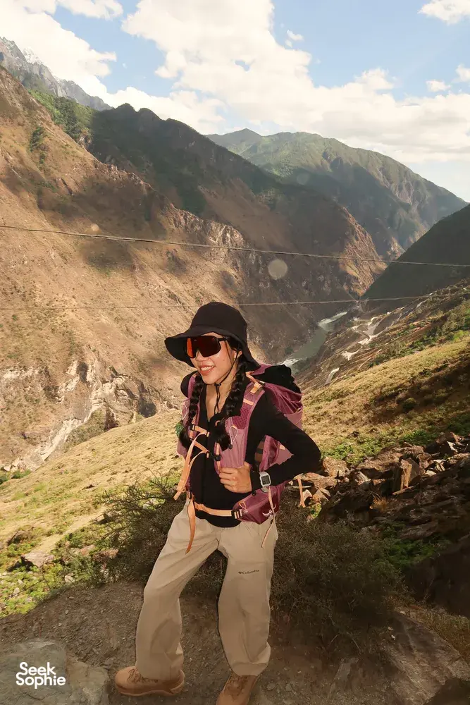 Hiker posing on a rocky ledge on the lesser-known high trail of Tiger Leaping Gorge in Lijiang, with The Bivou Lijiang