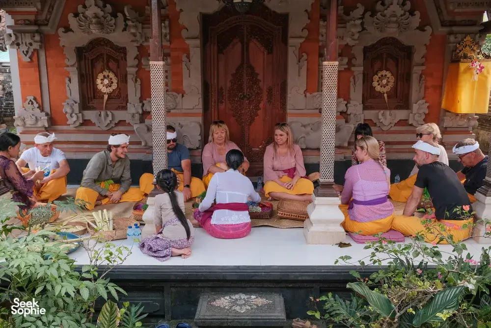 Group seated in a Balinese temple pavilion with canang sari offerings on the altar and cushions, Ubud