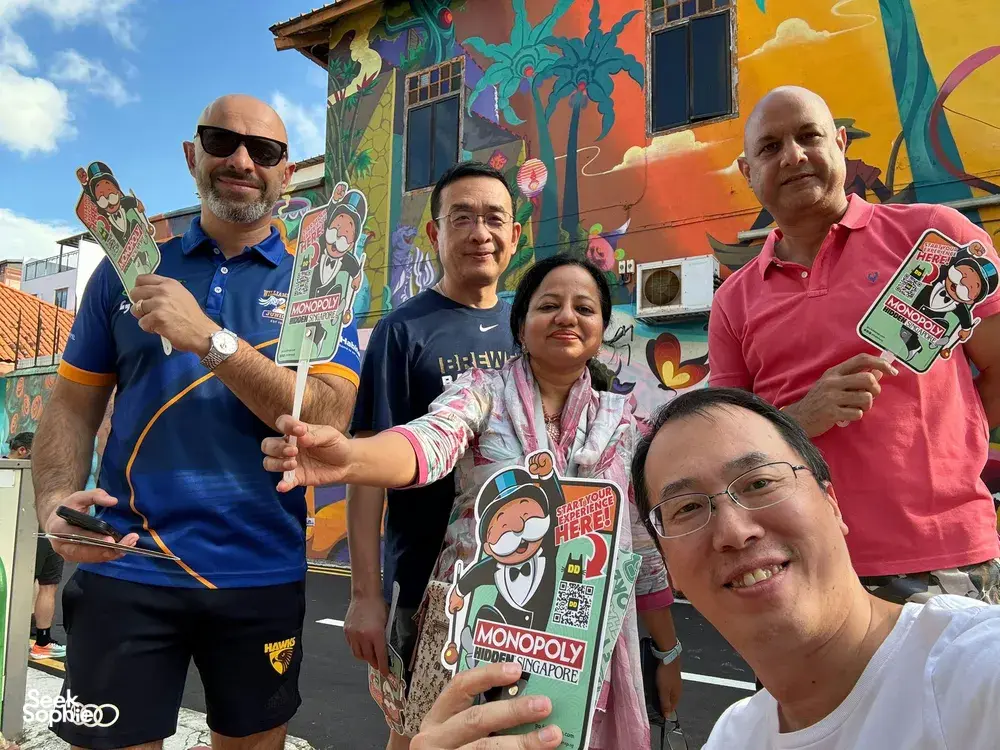 Team members holding Monopoly Hidden Singapore guides in front of a mural on Katong-Joo Chiat streets, for team building Singapore
