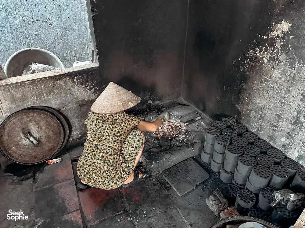Conical-hatted cook tending a smoky stove in a Hanoi home kitchen, where tofu is prepared traditionally