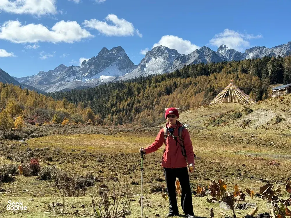 Hiker in a red jacket stands on a grassy trail with distant snow peaks and forested slopes in Shangri-La during the trek