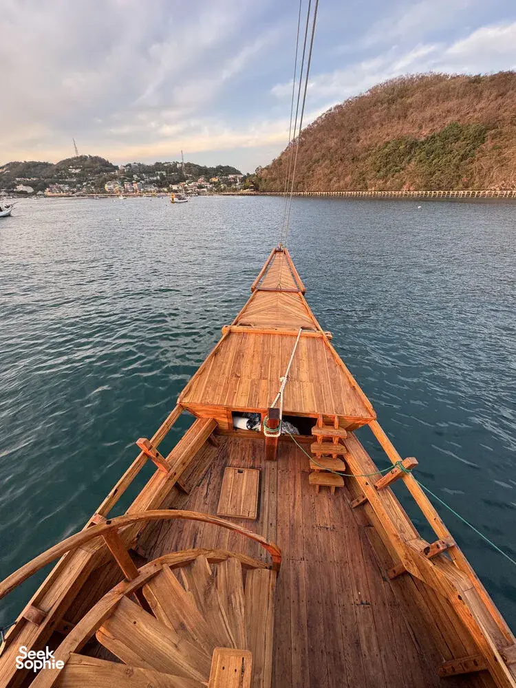 Bow of a wooden phinisi steering through calm water with hills and harbour boats at dusk near Labuan Bajo