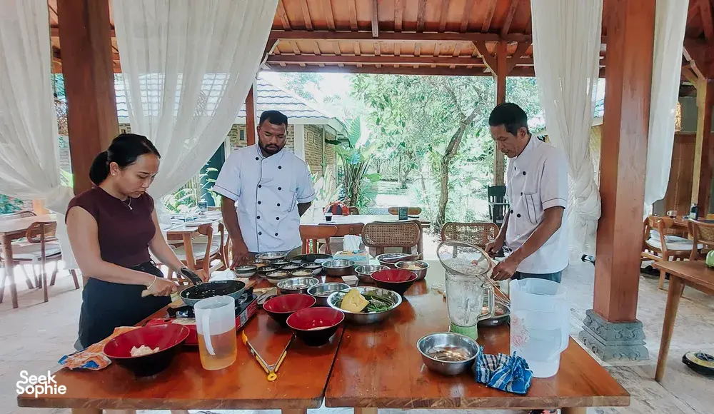 Small group cooking around pans and bowls, learning vegan Manggarai dishes from a local chef during a class in Labuan Bajo