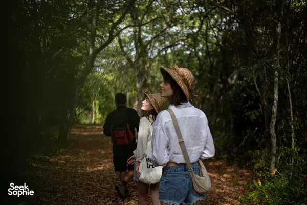 Woman walking along a forest trail in Indonesia during a retreat, carrying a backpack and drinking bottle behind her