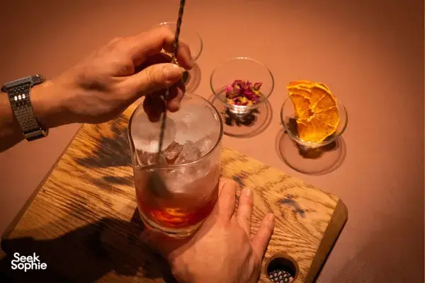 Mixologist pours chilled ingredients into a large glass on a wooden board in a Singapore cocktail-making session.