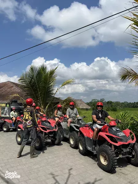 Helmeted riders on red ATV quad bikes lined up on a sunny Bali lane, with palms and green fields beyond