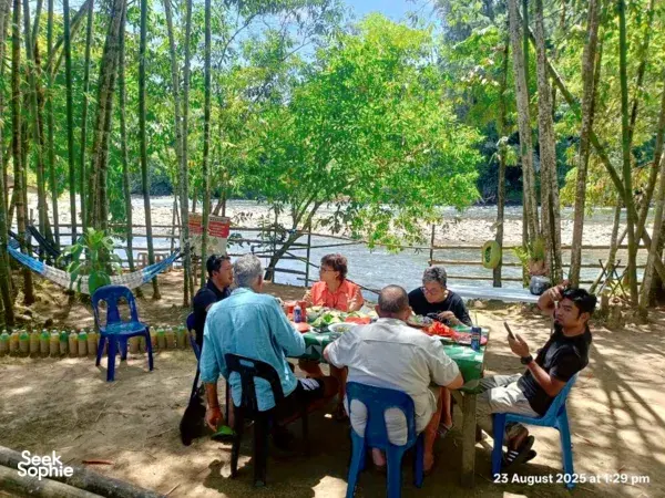 Friends sharing lunch at a shaded riverside table beneath tall trees, during Outreach Borneo’s rafting and survival day in Sabah