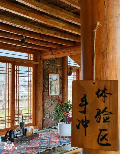 Rustic wooden tea room interior with large pinned calligraphy panel, stone wall and windows, with cups and kettle on a table in Lijiang