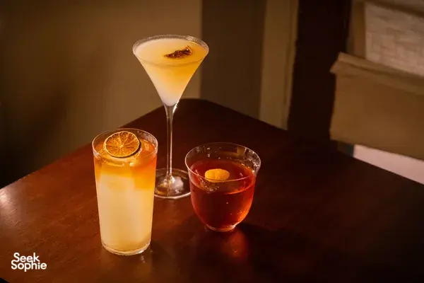Three citrus cocktails on a wooden tabletop in a cosy Singapore cocktail bar, each garnished with an orange slice.