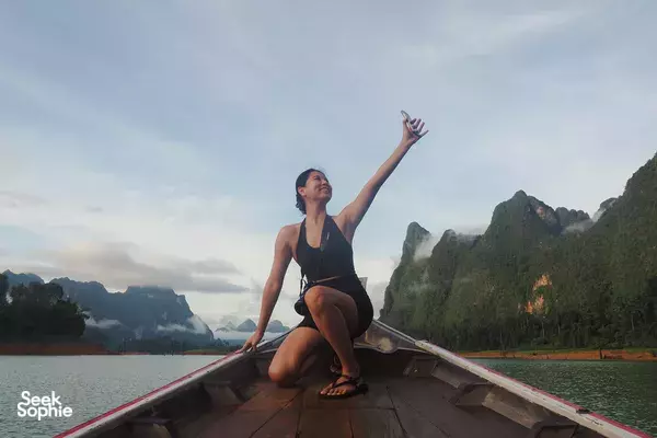 Woman kneeling on a wooden raft, waving at misty limestone karsts rising from the water on Cheow Lan Lake