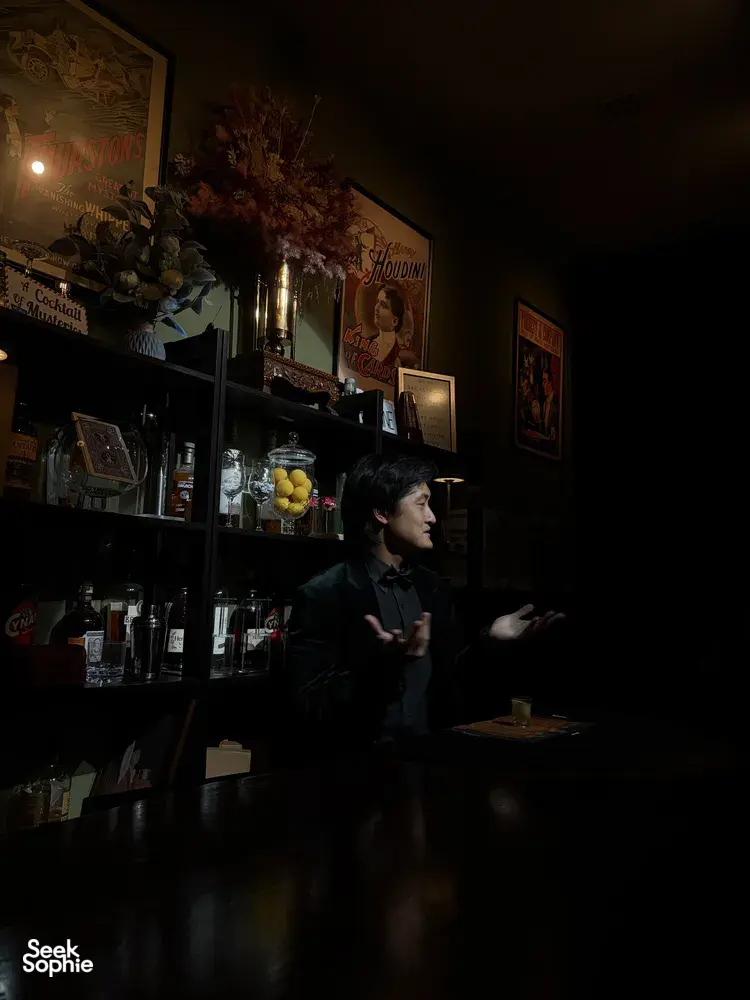 Seorang penghibur membuat isyarat dari belakang kaunter bar di The Magic Bar, Singapura, dengan pencahayaan malap dan botol-botol kaca di sekelilingnya