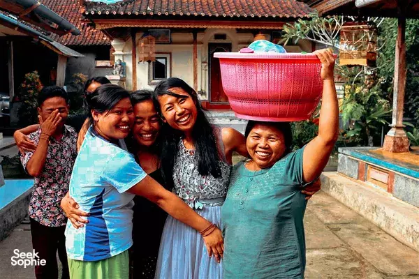 Group of women laughing in a sunny Indonesian courtyard, one holding a large woven pink basket overhead, surrounded by traditional buildings