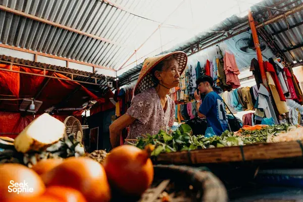 Local market trader in a patterned conical hat arranging greens at a colourful stall in Hoi An for home cooking