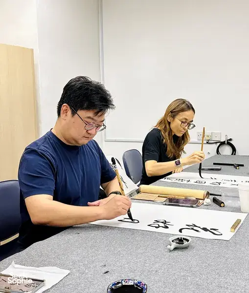 Chinese calligraphy workshop in Singapore with two learners writing on rice paper at tables, brushes and ink ready