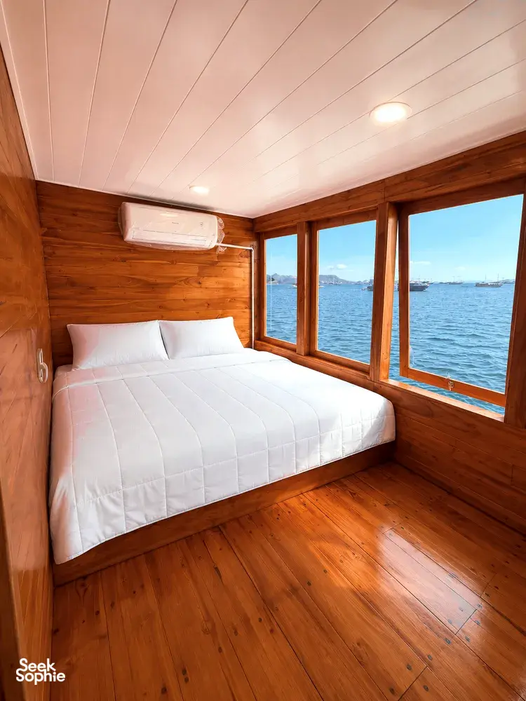 Wooden phinisi bedroom with double bed, bedside lights and sea-view windows on La Famille’s 3D2N shared Komodo cruise from Labuan Bajo