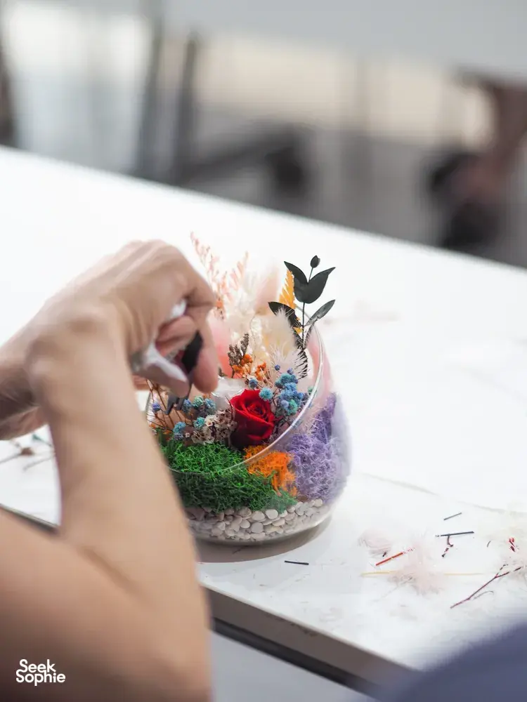 Hand placing preserved flowers into a glass floral bowl terrarium on a white tabletop during a Floral Bowl workshop in Singapore