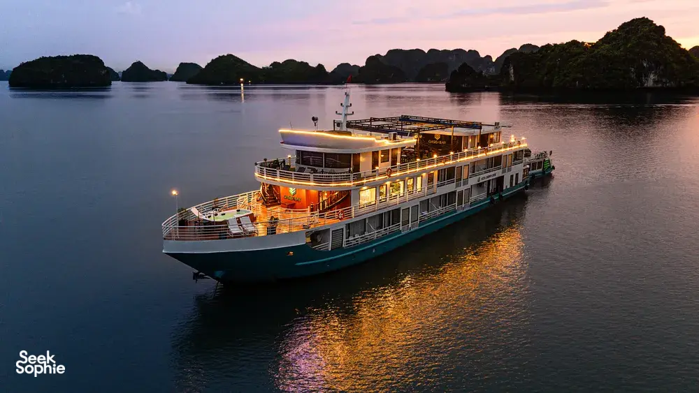 Oasis Bay party cruiser at dusk on Halong Bay with warm deck lights and silhouetted karst islands reflected in the water