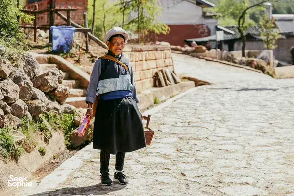 Traditional-dressed local woman standing on a stone path in Dali, Yunnan, holding supplies during a private car journey