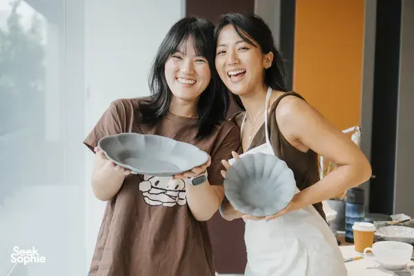 Two women smiling and holding painted ceramic plates and bowls at an Ina Atelier pottery workshop in Singapore