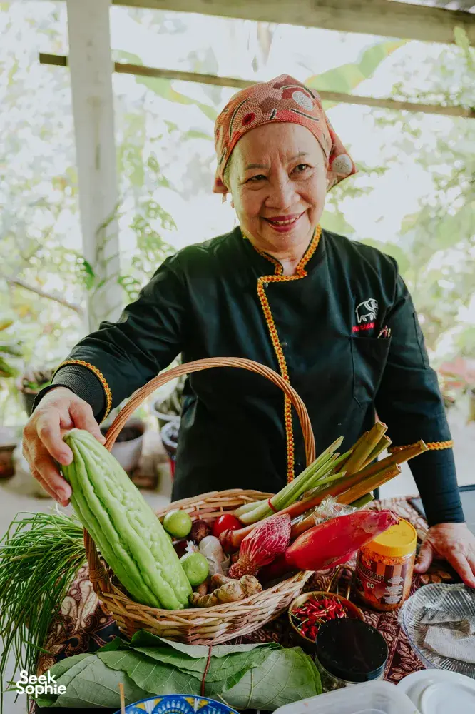 Indigenous Sabahan cook in Kota Kinabalu holding a basket of fresh greens and herbs, wearing traditional headscarf and dress