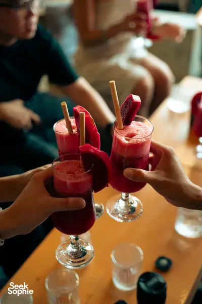 Hands clinking red fruit cocktails with melon wedges and paper straws at a table during a Hoi An cocktail tour