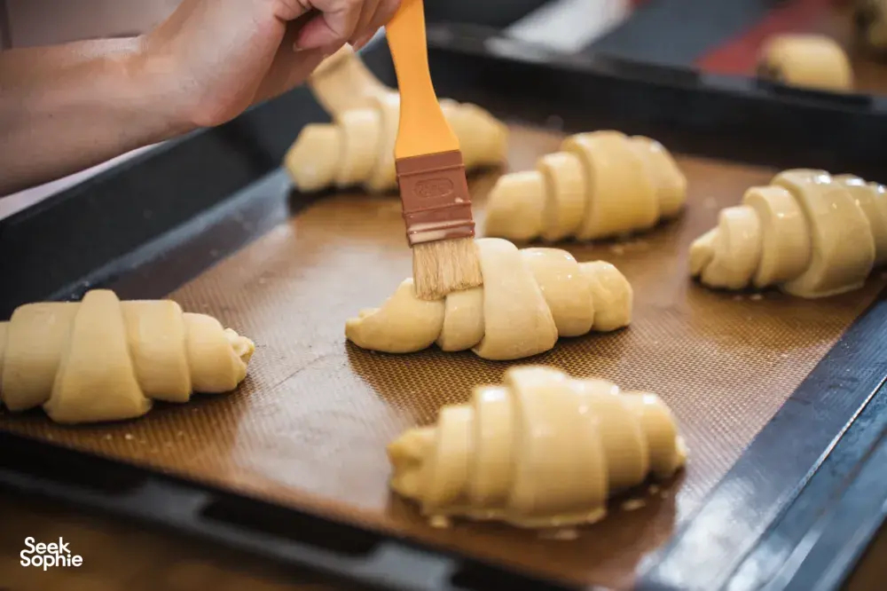 Chef brushing egg wash onto rolled croissant dough on a mesh-lined tray at French Cooking Lab’s Singapore class