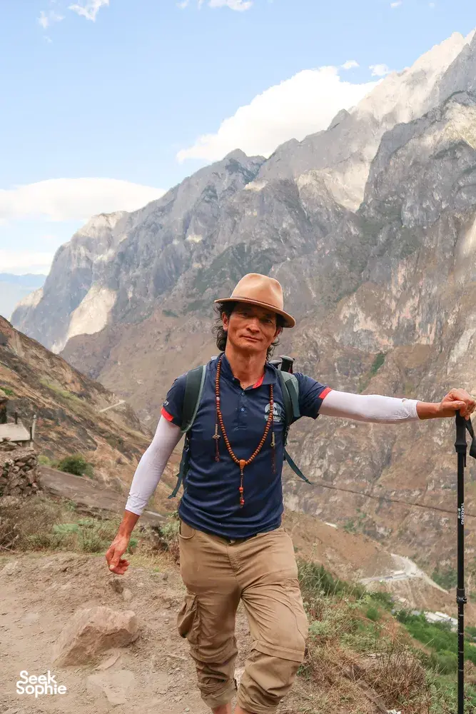 Trek leader in a wide-brim hat guiding hikers along a mountain slope on the high trail of Tiger Leaping Gorge in Lijiang, with The Bivou Lijiang
